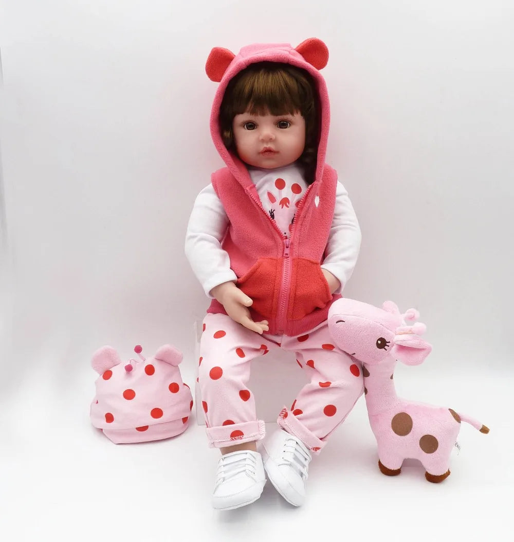 A baby-like reborn doll wearing a pink hooded outfit and pink pants, sitting on a white background.