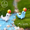 Two miniature animal figurines with colorful heads are placed on a mossy ground beside a blue stream.