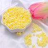 A white dish filled with yellow pearl beads and loose yellow pearl beads on a white satin cloth next to a pink rose.