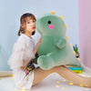 A young woman is sitting next to a large, soft green dinosaur plush toy on a white surface.