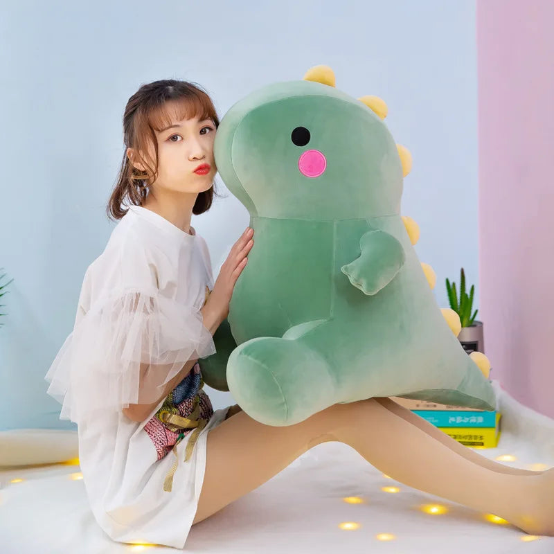 A young woman is sitting next to a large, soft green dinosaur plush toy on a white surface.