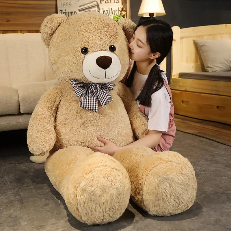 A young woman is sitting on the floor with a large tan teddy bear plush toy and a pink shirt in a wooden room.