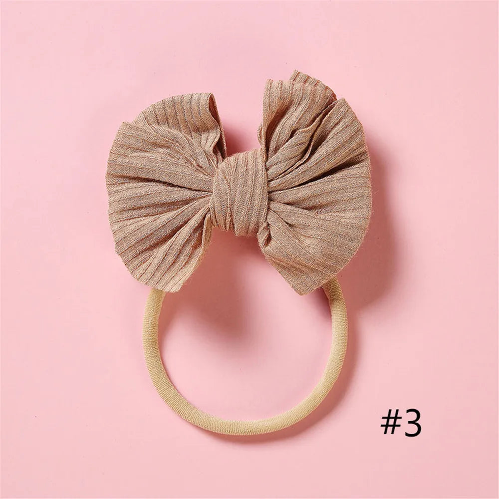 A beige fabric bow headband with a soft elastic band is shown against a pink background.