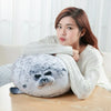 A woman is leaning on a large, round, white and gray seal plush pillow indoors.