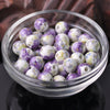 A clear glass bowl filled with purple and white floral ceramic beads for jewelry making