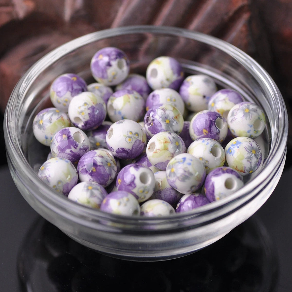 A clear glass bowl filled with purple and white floral ceramic beads for jewelry making