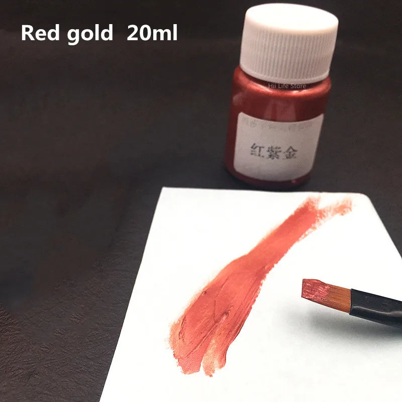 A small glass bottle of red gold acrylic paint is shown next to a swatch of the same reddish-orange color on white paper.