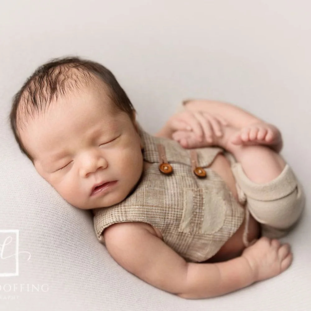 A newborn baby is lying down wearing a beige plaid vest and bundled in soft fabric, photographed in a studio setting.