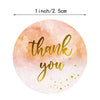 A round sticker with a soft peach watercolor background and gold script that reads thank you