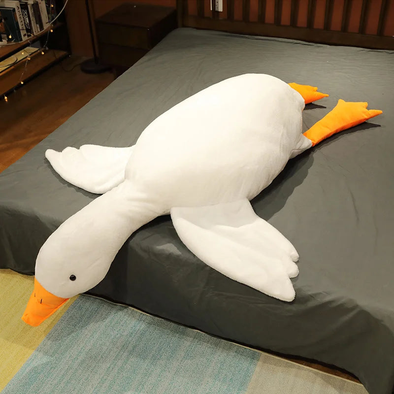 A white oversized kawaii goose plush pillow with orange accents resting on a gray bedsheet.