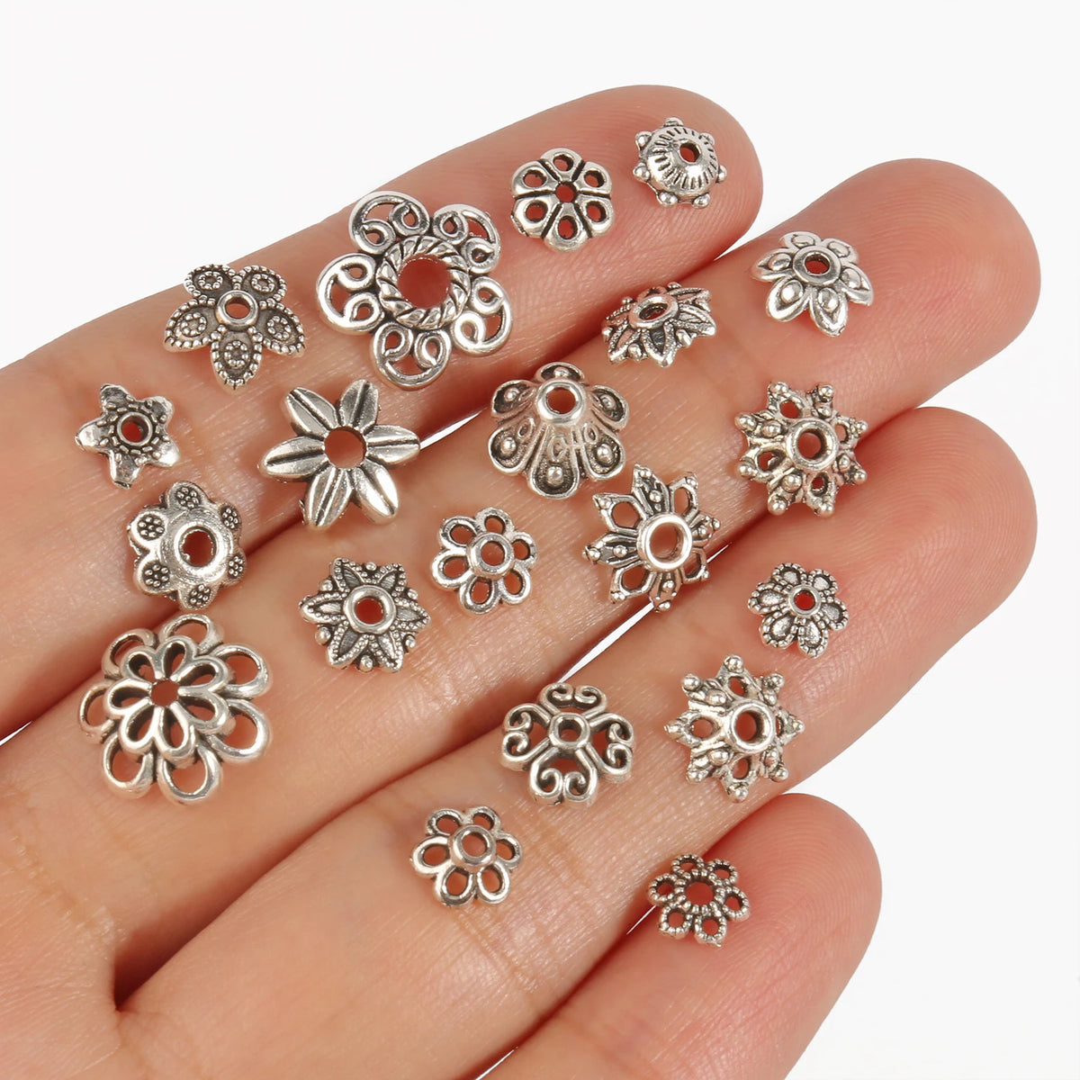 A hand holds various silver Tibetan style hollow flower bead caps for jewelry making.