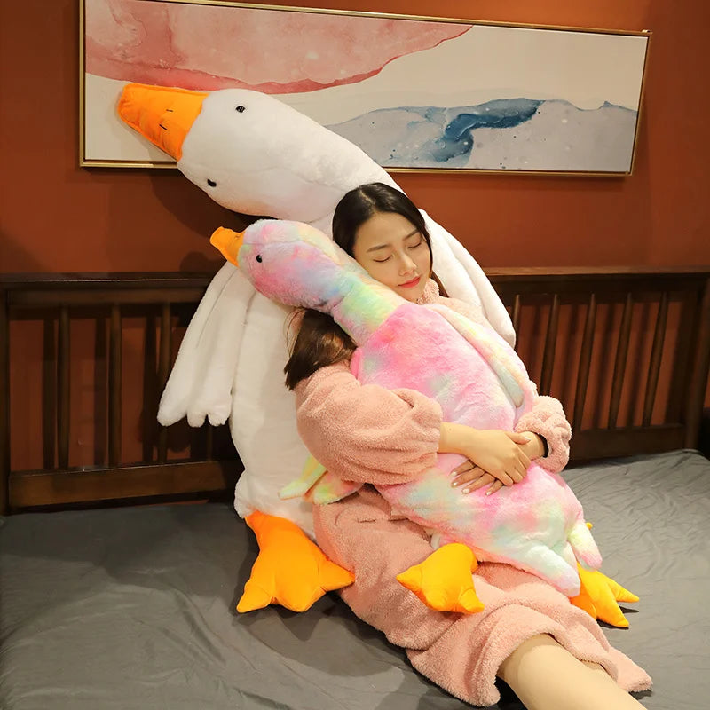 A woman is cuddling with an oversized white goose plush pillow and a pink and yellow goose plush pillow.