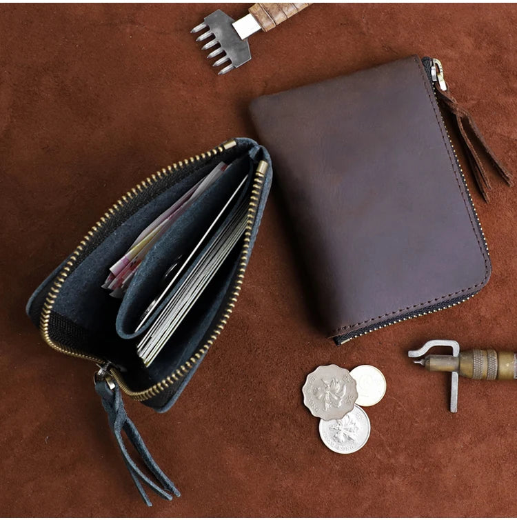 A dark brown leather coin purse with a zipper and a small leather tool are displayed on a brown surface.