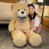 A young woman is sitting with a large, light brown teddy bear plush toy in a cozy indoor setting.
