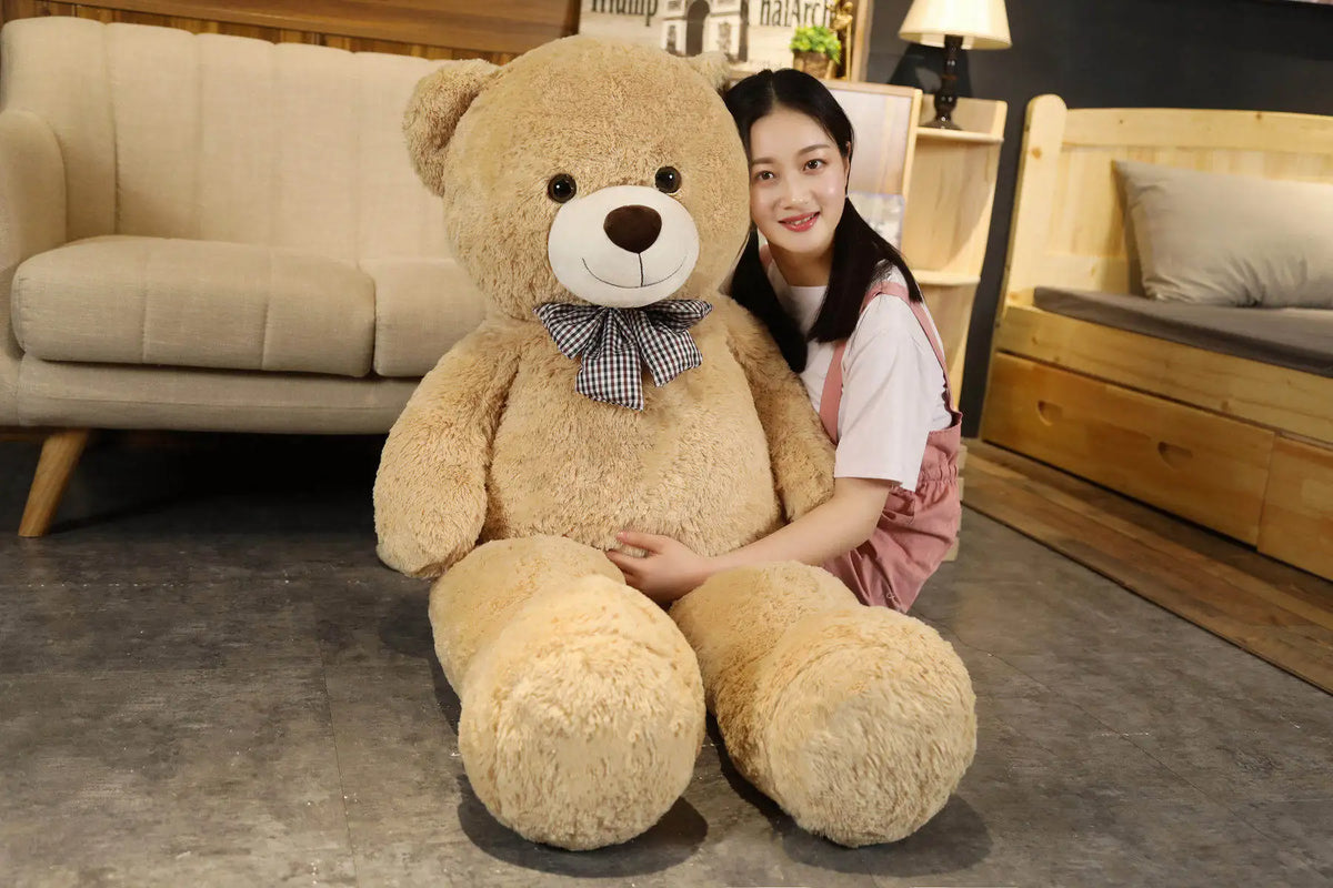 A young woman is sitting with a large, light brown teddy bear plush toy in a cozy indoor setting.