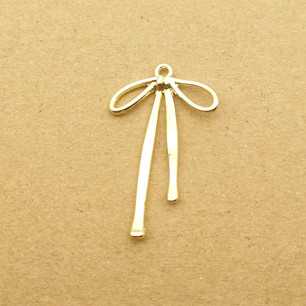 A gold-colored alloy bow charm with a loop and hanging strands is displayed on brown paper.