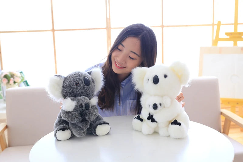 A young woman smiles while holding two fluffy white koala plush toys on a white table.