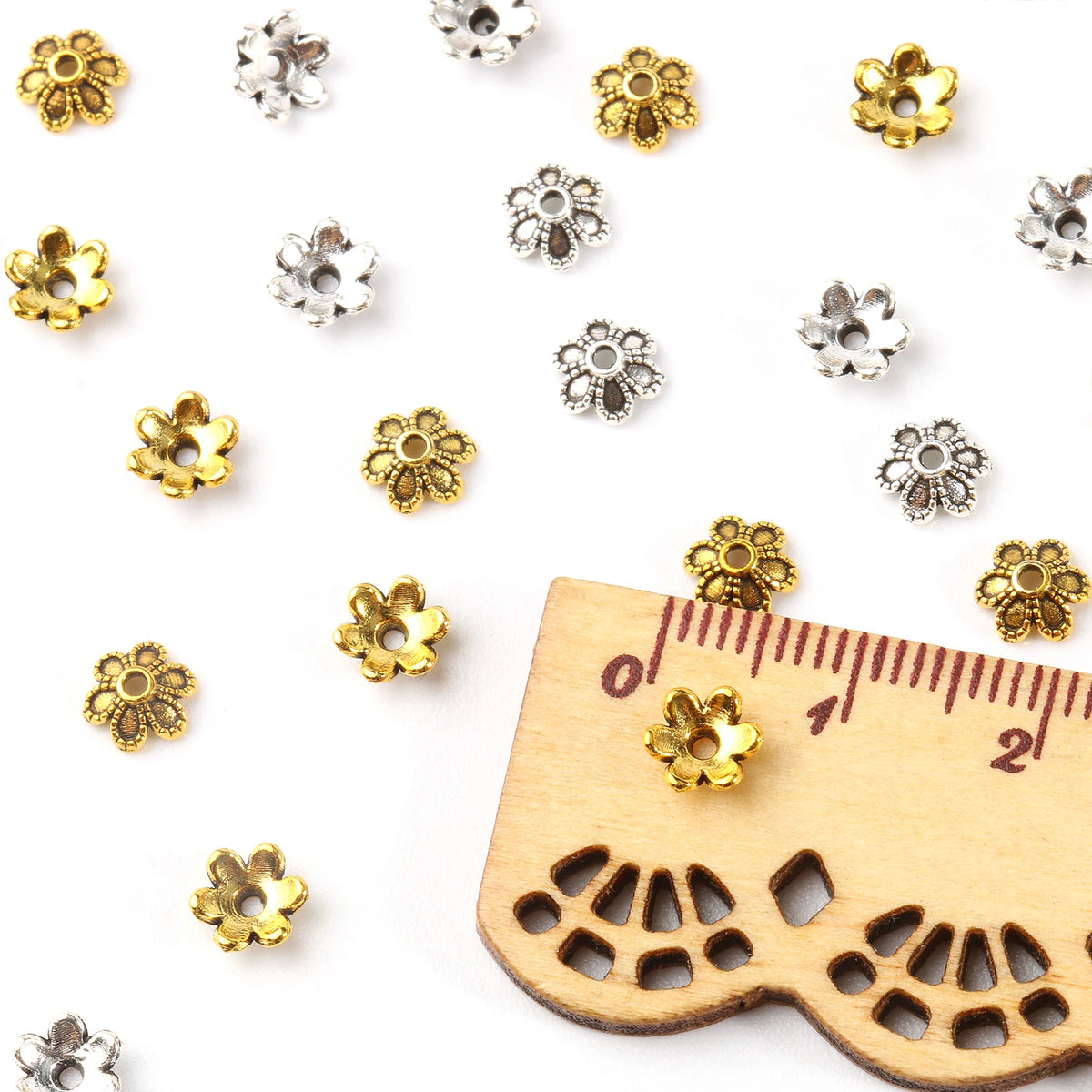 A collection of small silver and gold hollow flower bead caps for jewelry making displayed on a wooden ruler.