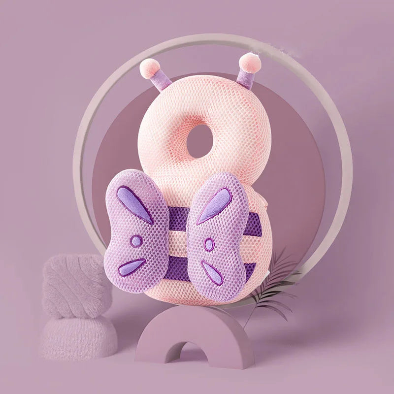 A pink soft baby head protector pillow with purple adjustable straps is displayed on a light purple surface.