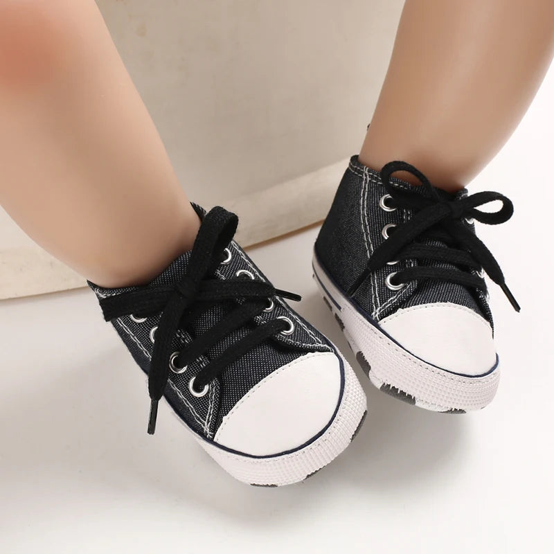 A pair of black canvas first walkers for infants shown on a person's foot.