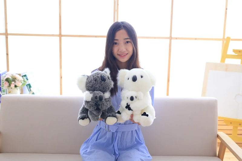 A young woman sits on a couch holding two plush koala toys, one gray and one white.