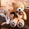 A smiling woman is posing with a large, tan plush teddy bear wearing a brown knit scarf and paw prints.