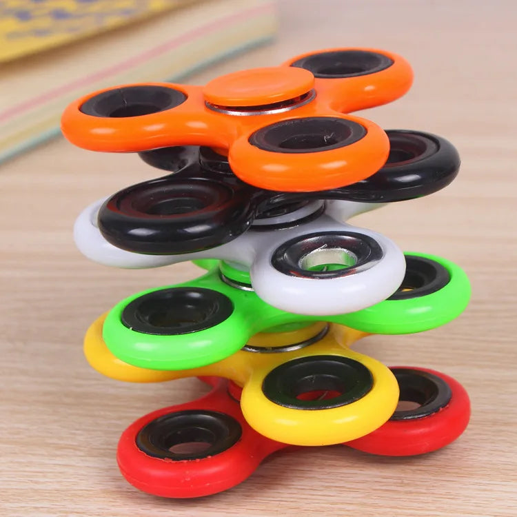 A stack of colorful ABS tri-spinner fidget toys in orange, black, white, green, yellow, and red on a wooden surface.