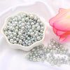 A white bowl filled with assorted silver and gray pearl beads, next to a pink lotus flower.
