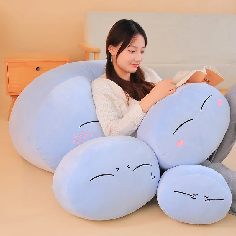 A light blue Rimuru Tempest plush toy pillow with a cute face is resting on a beige surface.