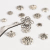 A silver tool is shown holding a detailed, openwork flower bead cap for jewelry making, surrounded by similar small silver be