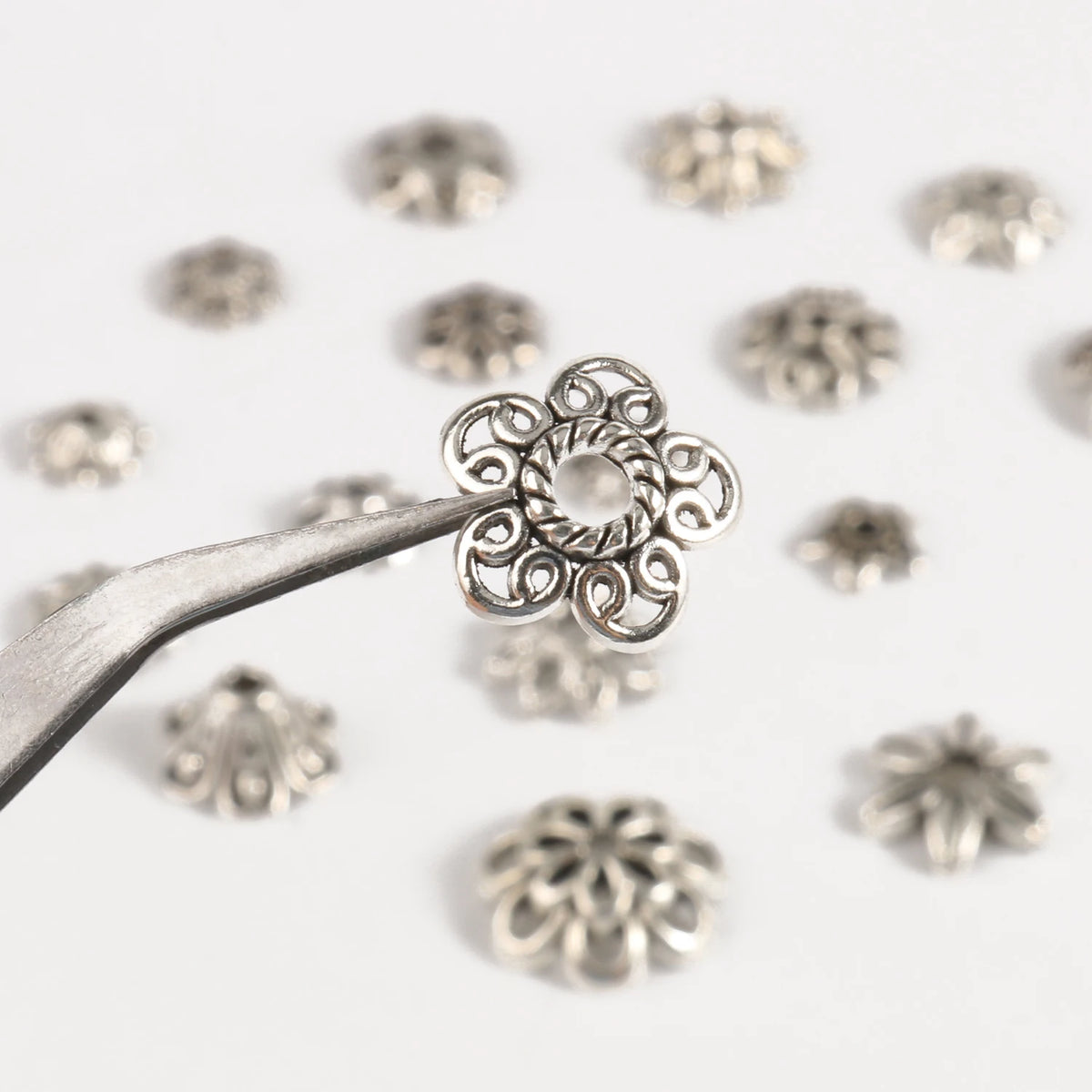 A silver tool is shown holding a detailed, openwork flower bead cap for jewelry making, surrounded by similar small silver be