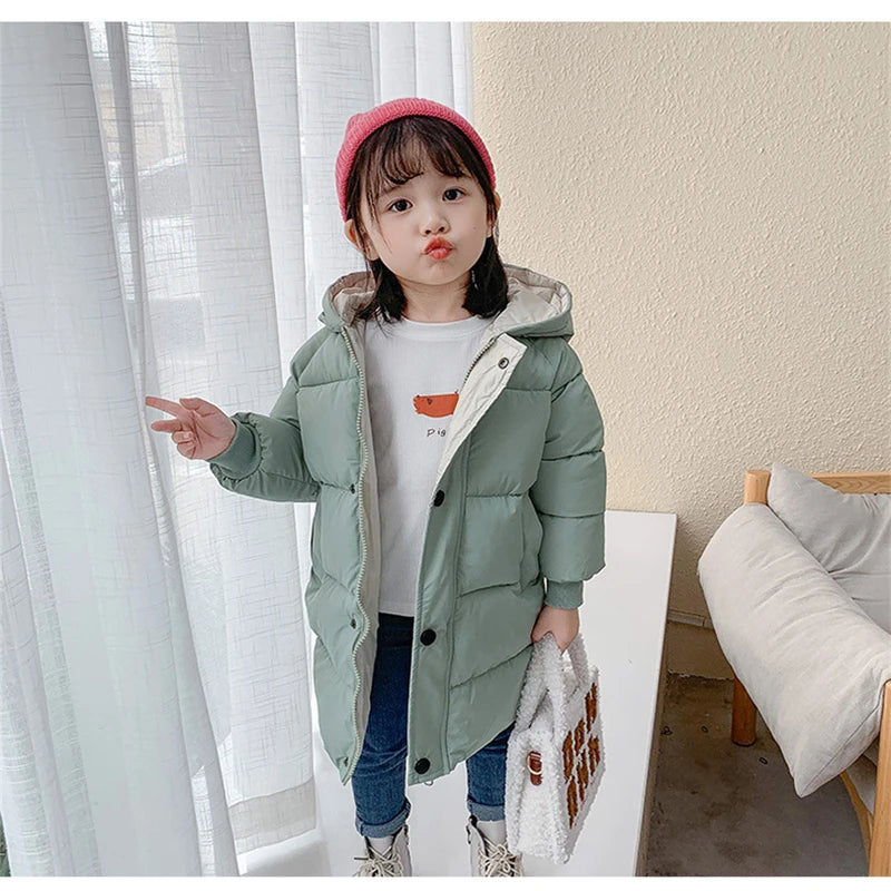 A young girl wearing a mint green padded parka jacket with a hood and a pink beanie standing indoors.