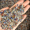 A hand holds a pile of natural mixed quartz crystal stones in various colors including clear, purple, blue, and reddish-brown