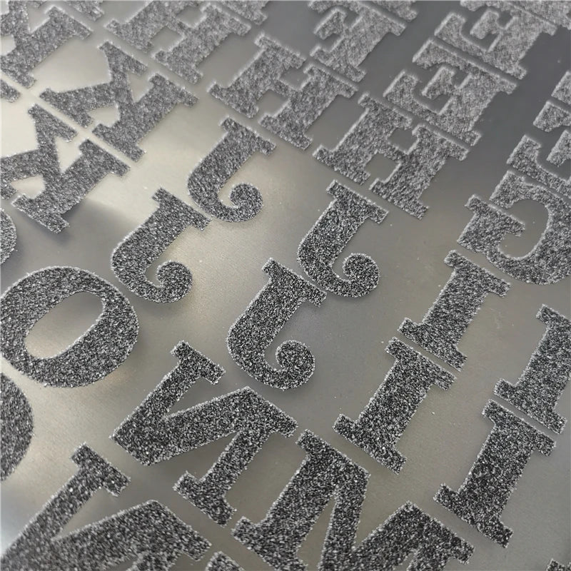 Close-up of silver glitter iron-on heat transfer patches with various letters and symbols on a gray surface.