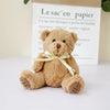 A soft brown patch bear plush toy with a yellow ribbon, displayed in front of a paper bag.
