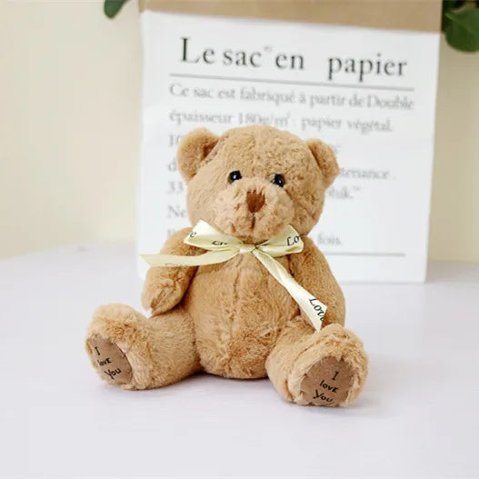 A soft brown patch bear plush toy with a yellow ribbon, displayed in front of a paper bag.