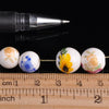 A row of white ceramic beads with floral designs is displayed next to a black pen and a wooden ruler.