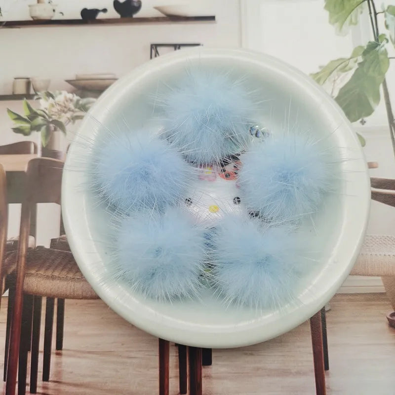 A close-up of light blue fluffy mink fur pom poms displayed in a white bowl on a wooden table.