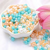 A bowl filled with assorted pearl beads in shades of pink, cream, and blue scattered on a white surface.