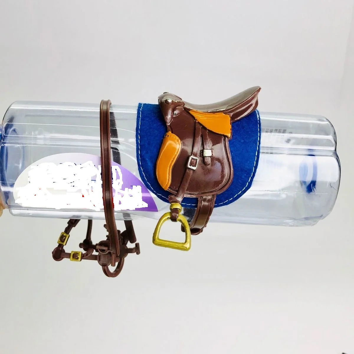 A brown horse saddle and riding prop accessory with a blue fabric backing is displayed on a clear plastic bottle.
