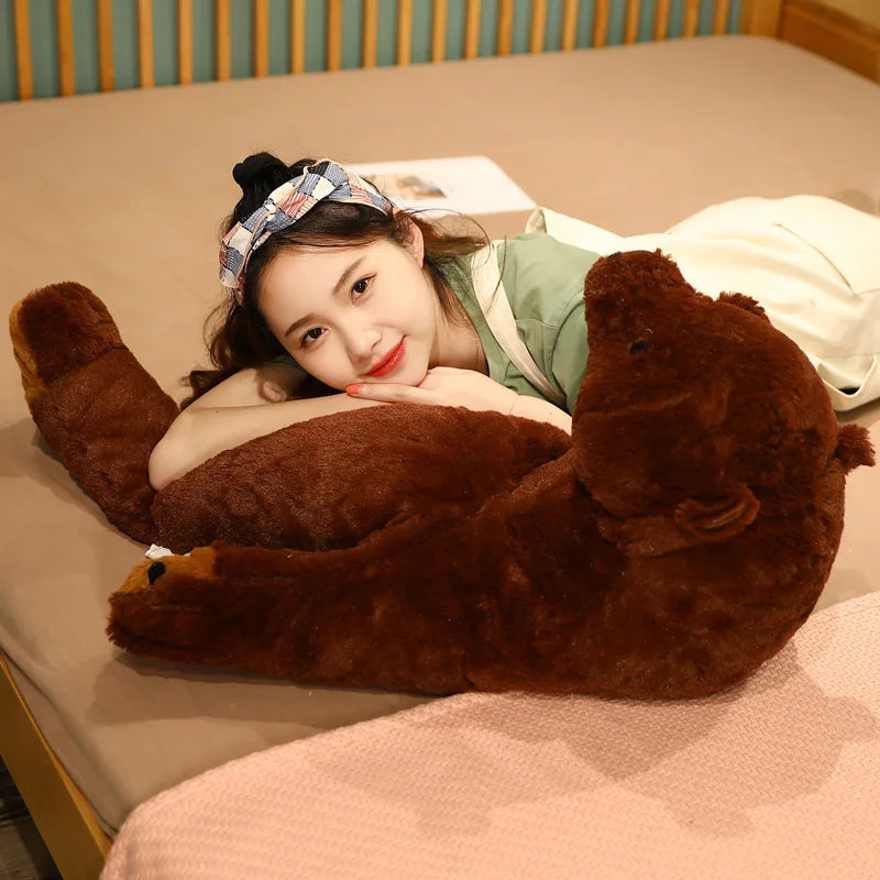 A young woman is lying down with her arms around an oversized lifelike brown bear plush cushion on a beige surface.