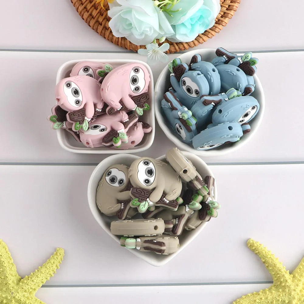 A collection of adorable sloth silicone beads in various shades of pink and blue are displayed on a white surface.