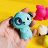 A hand holds a light blue animal finger puppet with large eyes and black ears against a yellow background.