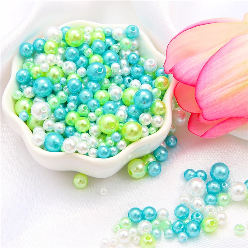 Assorted pearl beads in shades of blue and green are displayed next to a pink flower.