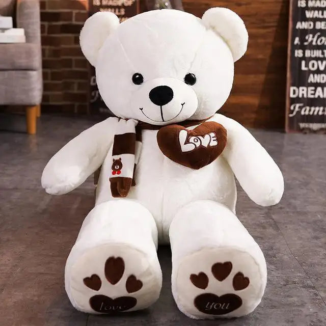 A white plush teddy bear wearing a brown heart-shaped accessory and knit scarf sitting on a gray floor.