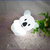 A white koala LED night light is illuminated on a sheer fabric surface with scattered gold stars.