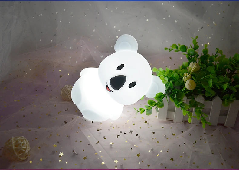 A white koala LED night light is illuminated on a sheer fabric surface with scattered gold stars.