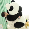A woman is hugging a large white and black panda plush toy indoors.