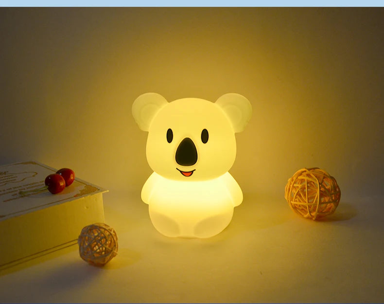 A glowing yellow koala night light sits on a surface next to a small wooden box and a spherical object.
