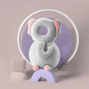 A soft gray elephant-shaped baby head protector pillow with pink ears is displayed inside a white circle against a purple bac